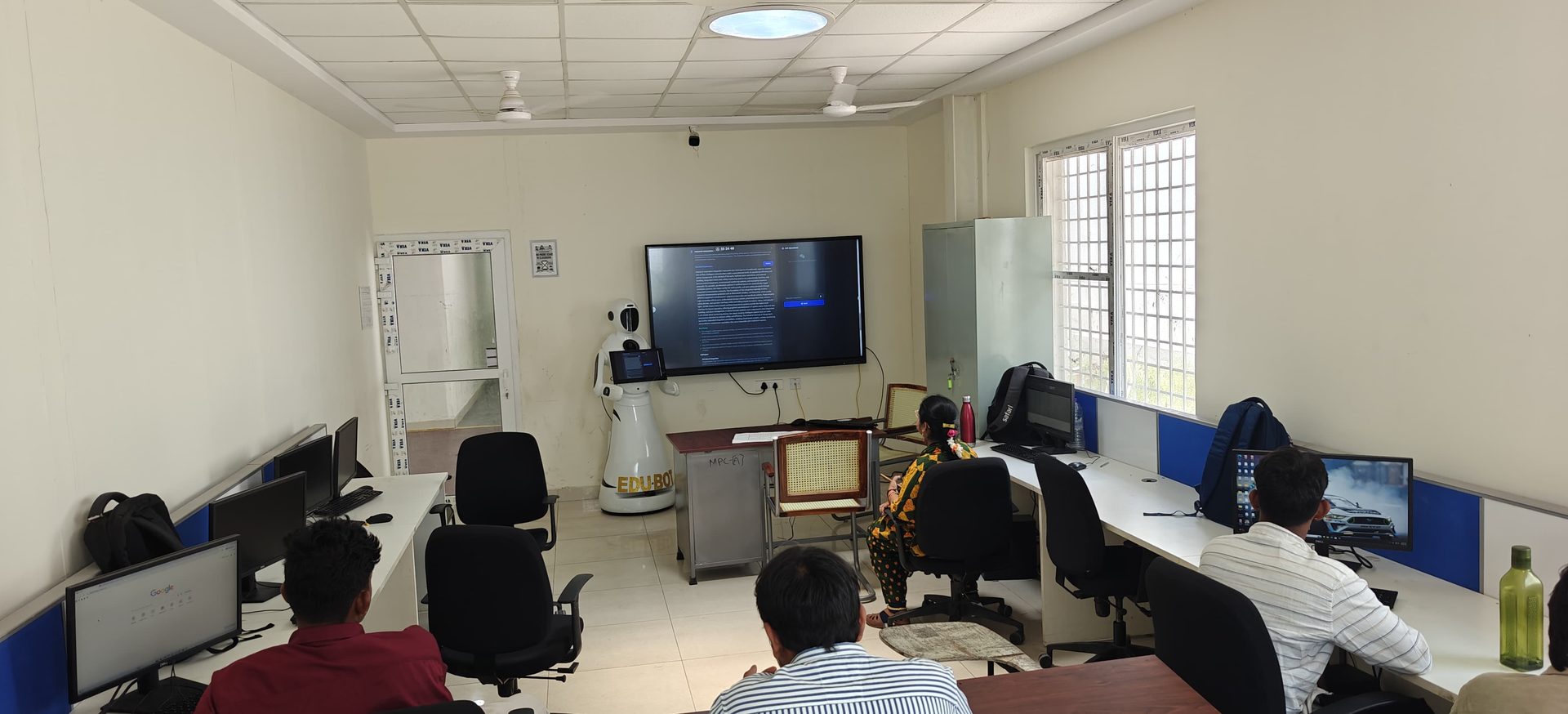 The robot standing at the front of a computer training lab while trainees work at desktop workstations