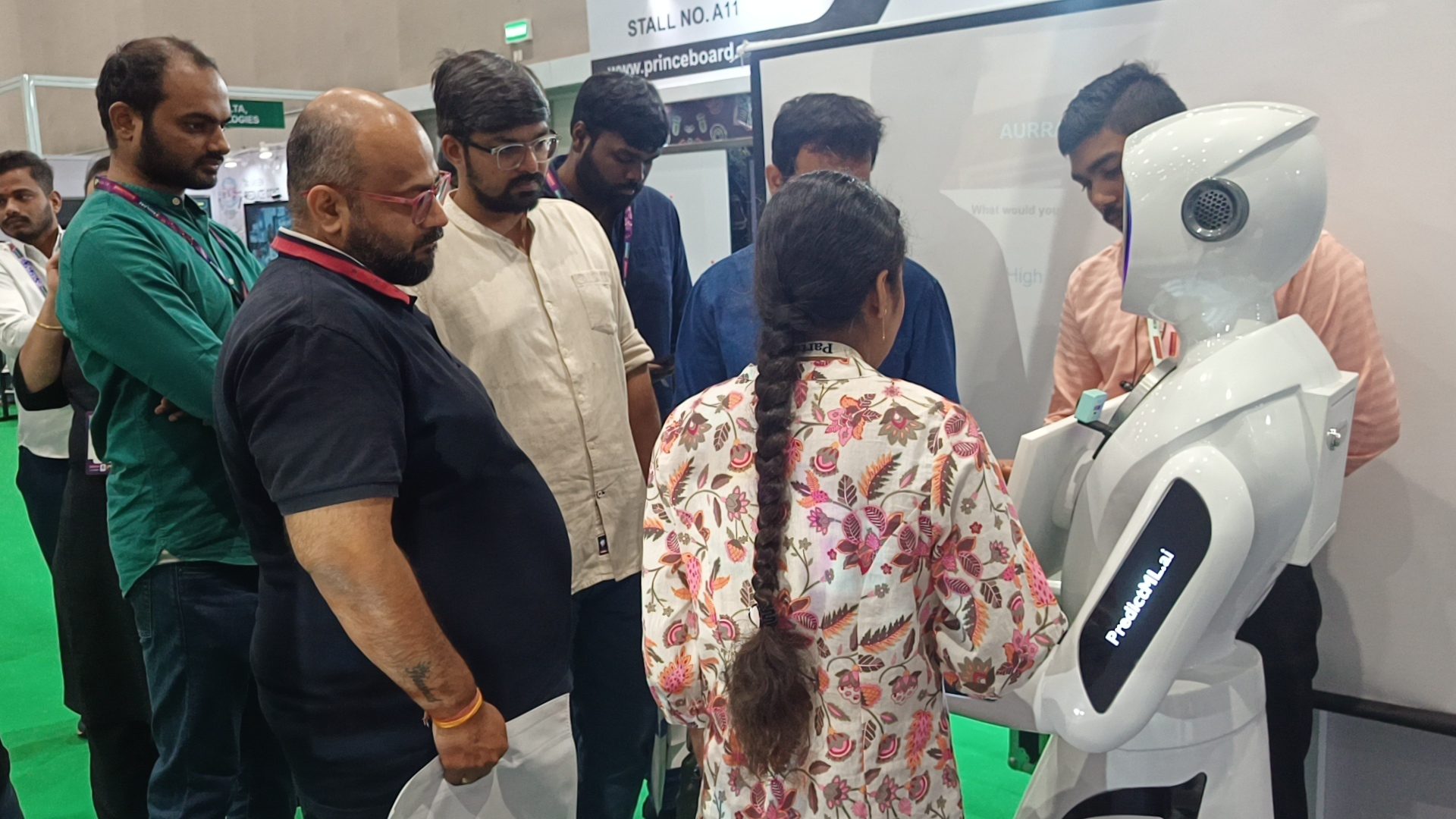 A group of exhibition visitors gathered around the robot, one reaching toward its touchscreen