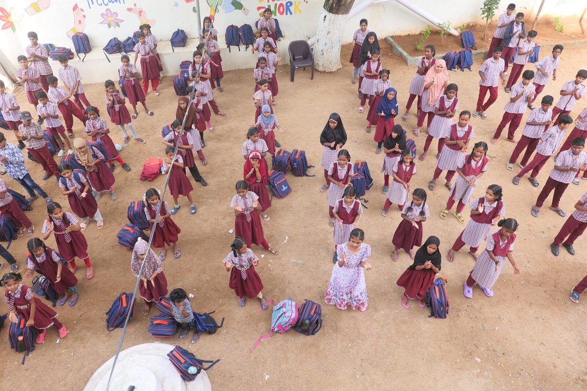 A school assembly in the yard, students standing in rows with their bags in front of them