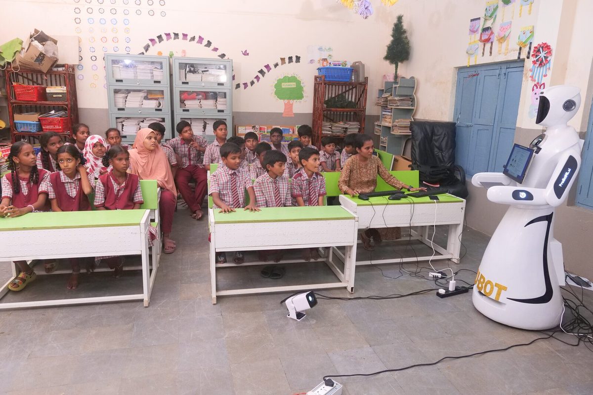 A government school classroom with students at green desks and the robot at the front of the room