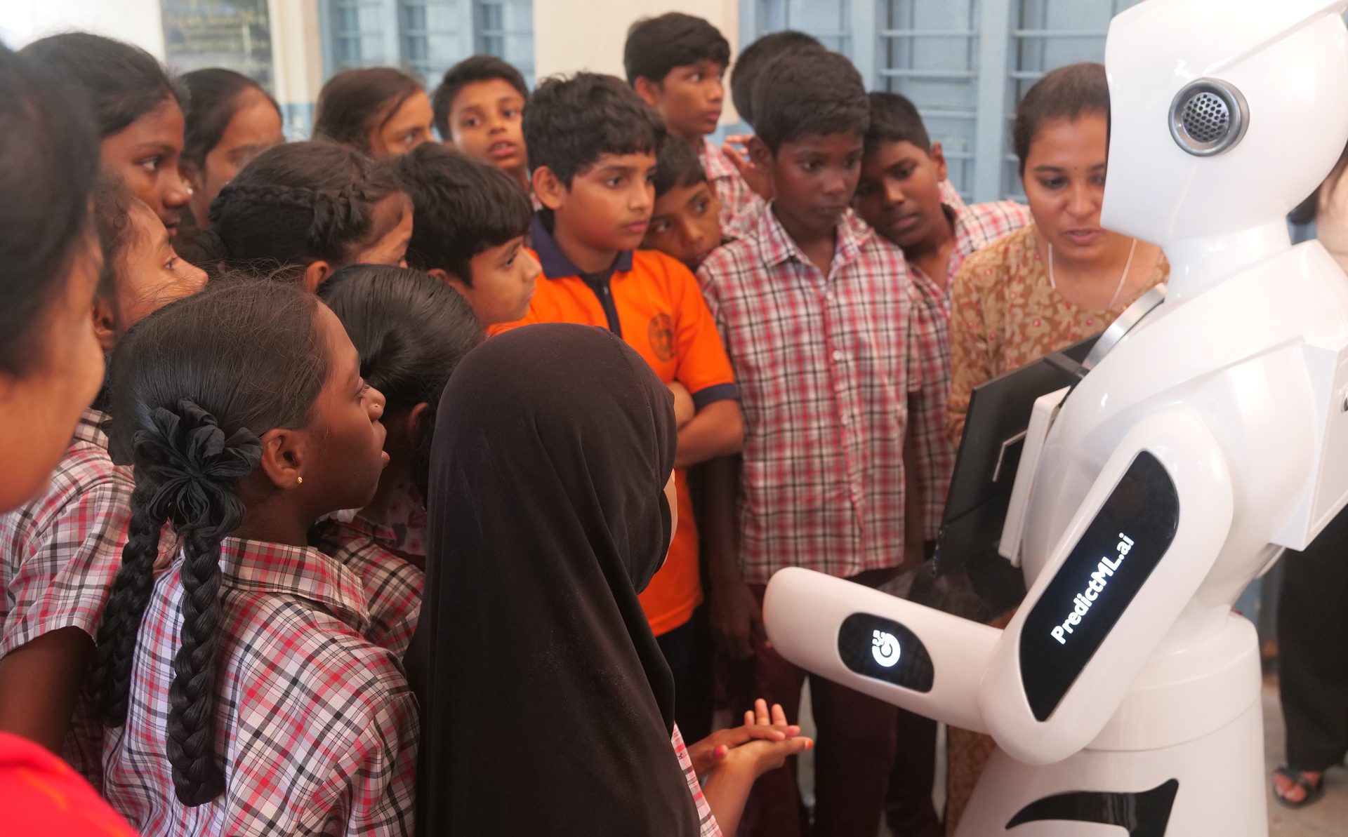 Children crowding closely around the robot with a teacher, several reaching toward its screen