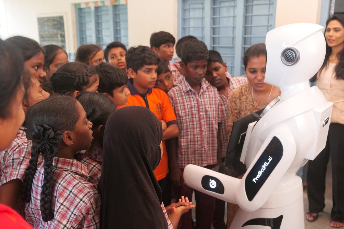 Students gathered around the robot in a school corridor, a teacher standing with them
